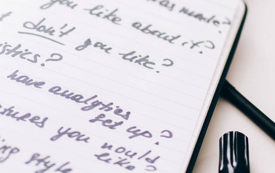 Notebook containing handwritten practice questions and study notes beside a pen, representing exam preparation for the CBSA Officer Trainee Entrance Examination (OTEE).