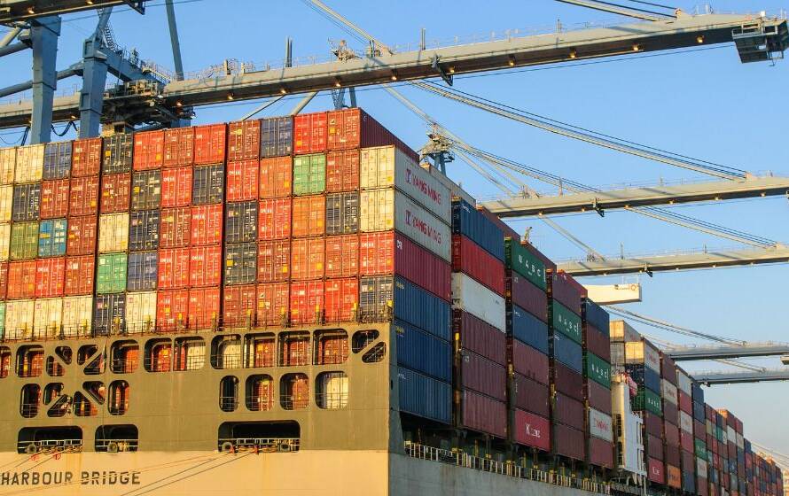 Container ship loaded with stacked cargo containers at a busy seaport, with large gantry cranes operating along the vessel.