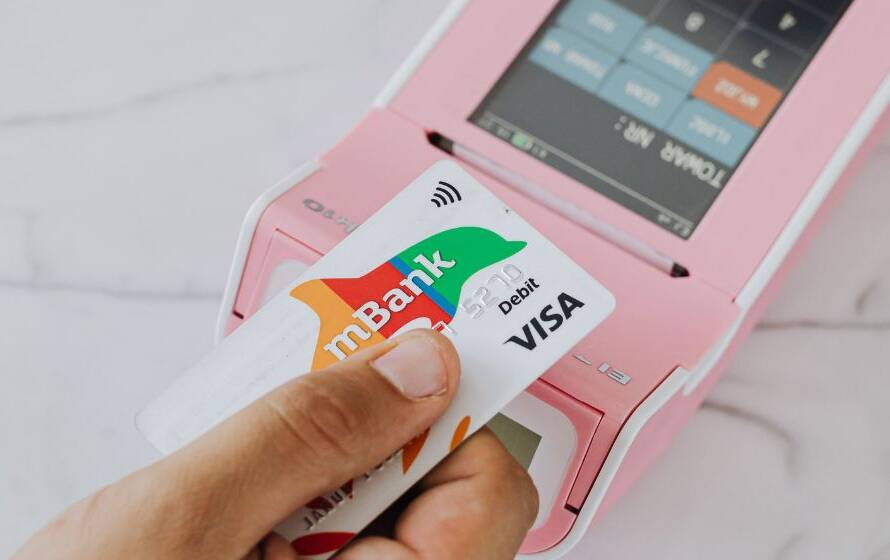 Hand using a debit card to make a contactless payment with a pink payment terminal, illustrating financial transactions and payroll deductions.